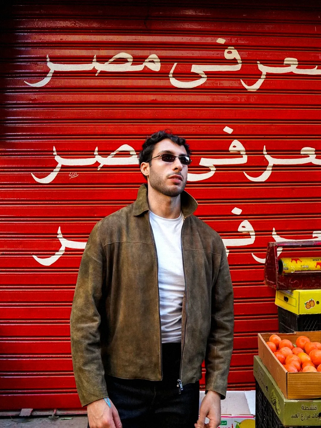 A young man in a jacket in front of a red shop shutter painted with Arabic lettering