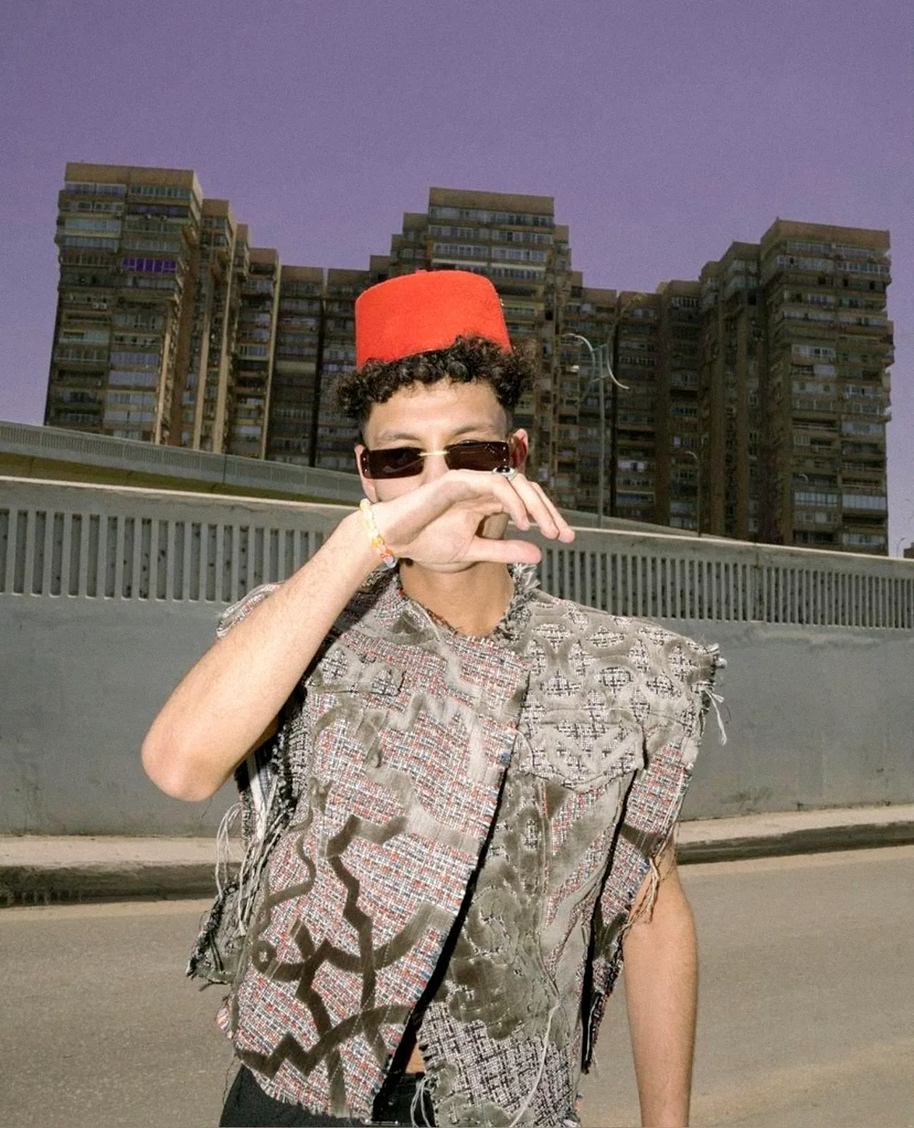 A young man in sunglasses and a red tarboosh making a peace sign in front of Cairo apartment blocks